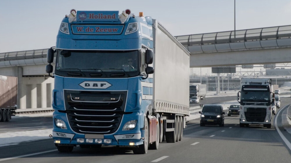 Na autostradzie jedzie niebieska ciężarówka z napisem \'Holland W. de Zeeuw\' oraz logo \'DAF\'. W tle widoczne są inne pojazdy oraz wiadukt.