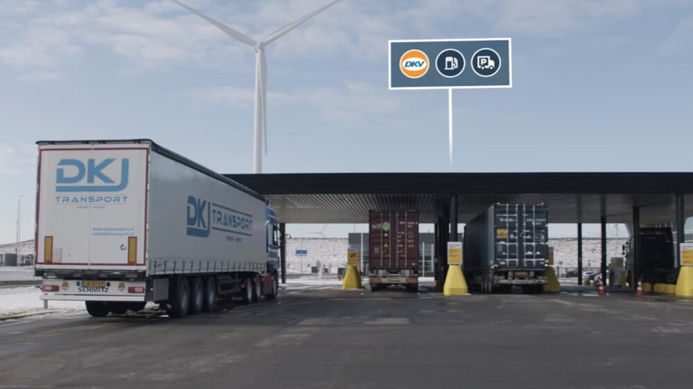 Na obrazie widać stację obsługi dla ciężarówek z zadaszeniem, pod którym znajdują się dwa pojazdy. W tle widoczna jest turbina wiatrowa, a na górze obrazu znajduje się znak z logo \'DKV\' oraz symbolami paliwa i parkingu.