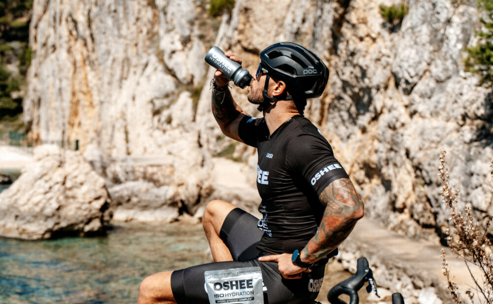 Izotonik OSHEE Hydration Zero Wieloowocowy (630 g) góry osoba w stroju sportowym i kasku pije izotonik z bidonu Hydro Synergy Complex™ OSHEE elektrolity wspierające prawidłową równowagę wodno-elektrolitową organizmu potas (2000 mg) magnez (400 mg) cynk (10 mg) w postaci diglicynianu cynku pomagają utrzymać prawidłową równowagę elektrolitową organizmu