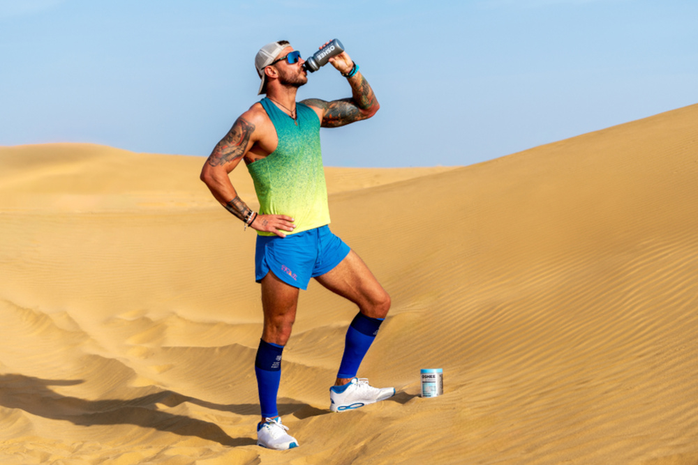 Izotonik OSHEE Hydration Zero Grejpfrutowy (330 g) piasek pustynia osoba pije napój z bidonu obok stoi opakowanie izotoniku odpowiedni poziom nawodnienia każdego dnia 22 porcje napoju lekka, grejpfrutowa formuła bez dodatku cukru doskonale orzeźwia wspiera organizm podczas aktywności fizycznej oraz w ciągu dnia jedna miarka produktu dostarcza składników wspierających nawodnienie oraz gospodarkę elektrolitową