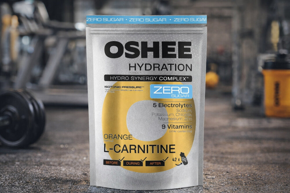 Izotonik OSHEE Hydration Zero Pomarańczowy (630 g) siłownia lekko rozmazane tło opakowanie izotoniku stoi na podłodze nawodnienie lekka formuła aktywność