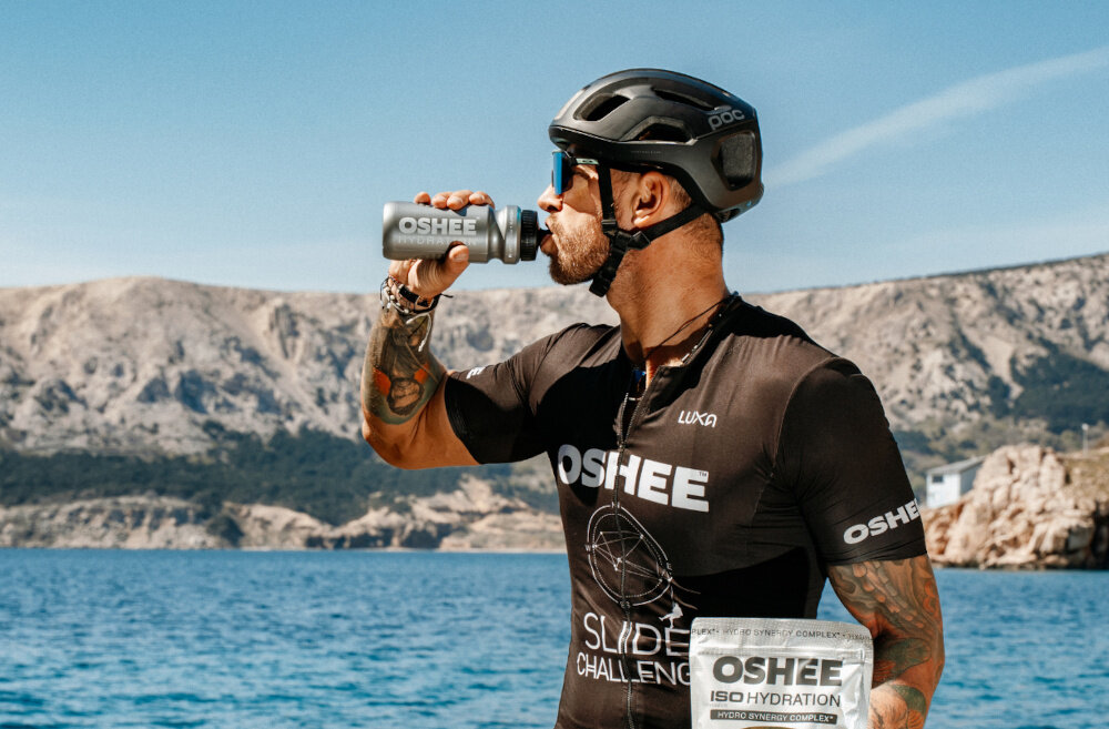 Izotonik OSHEE Hydration Zero Pomarańczowy (630 g) góry osoba w stroju sportowym i kasku pije izotonik z bidonu odpowiedni poziom nawodnienia każdego dnia 42 porcje napoju lekka, pomarańczowa formuła bez dodatku cukru doskonale orzeźwia wspiera organizm podczas aktywności fizycznej oraz w ciągu dnia jedna miarka produktu dostarcza składników wspierających nawodnienie oraz gospodarkę elektrolitową