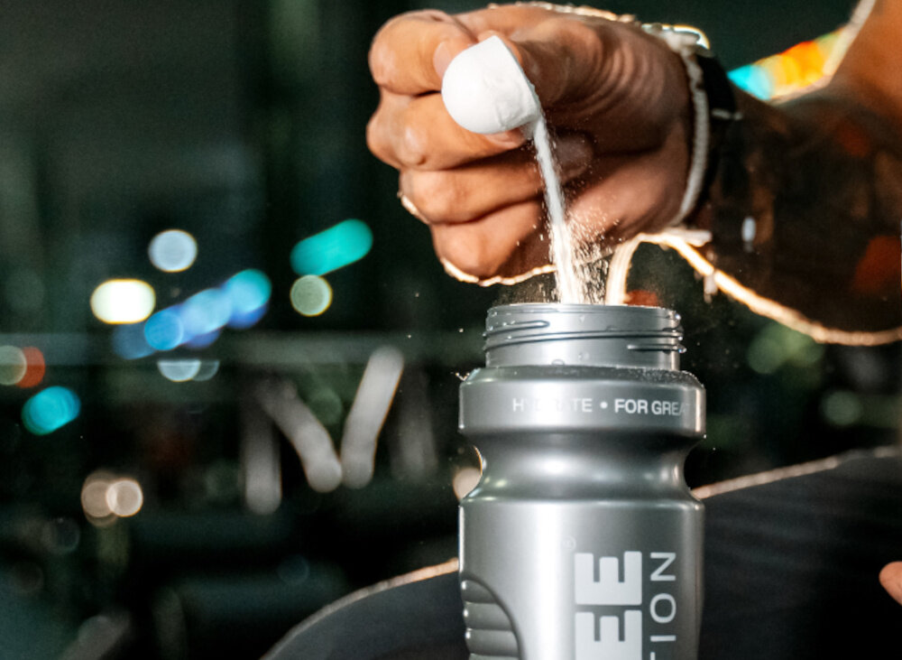 Izotonik OSHEE Hydration Zero Pomarańczowy (630 g) rozmazane tło osoba wsypuje miarkę izotoniku do bidonu do treningów na siłowni, biegania jazdy na rowerze treningów outdoorowych proszkowa forma specjalna miarka Jedną porcję 15 g proszku z 500 ml wody