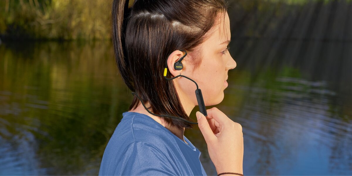 Słuchawki dokanałowe HAMA Freedom Athletics III Czarny Kobieta z profilu podczas treningu na zewnątrz, mająca założone czarne sportowe słuchawki Hama z neonowo-żółtymi akcentami. Słuchawki połączone są kablem biegnącym z tyłu głowy, a palce kobiety spoczywają na pilocie sterującym. W tle rozmyta tafla wody i zieleń.