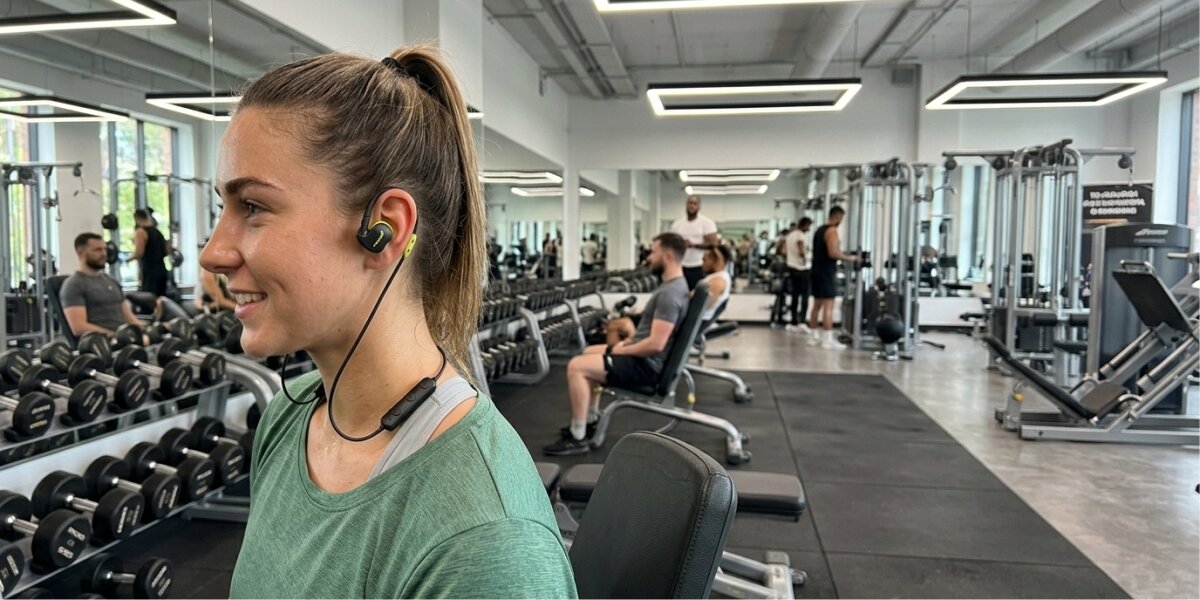 Słuchawki dokanałowe HAMA Freedom Athletics III Czarny Uśmiechnięta młoda kobieta trenująca na siłowni, korzystająca z czarno-żółtych słuchawek sportowych Hama. Słuchawki pewnie trzymają się na uszach dzięki pałąkom, a kabel z pilotem swobodnie zwisa na szyi. W tle widoczne nowoczesne wyposażenie siłowni, hantle i inni ćwiczący.