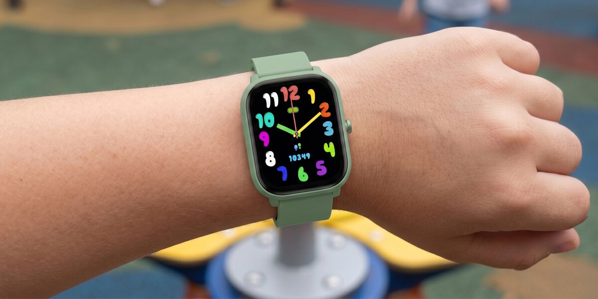 Osoba nosi zielony smartwatch z kolorowym wyświetlaczem, na którym widoczne są duże, różnokolorowe cyfry oraz liczba 10349. W tle znajduje się rozmyte otoczenie z elementami placu zabaw.