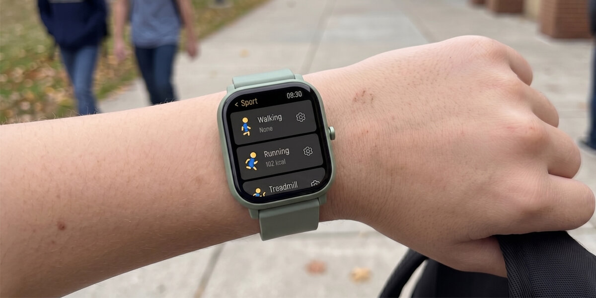 Osoba nosi zielony smartwatch z wyświetlaczem pokazującym aplikację sportową z opcjami: Walking, Running (102 kcal) i Treadmill. W tle widać chodnik i dwie osoby idące w oddali.