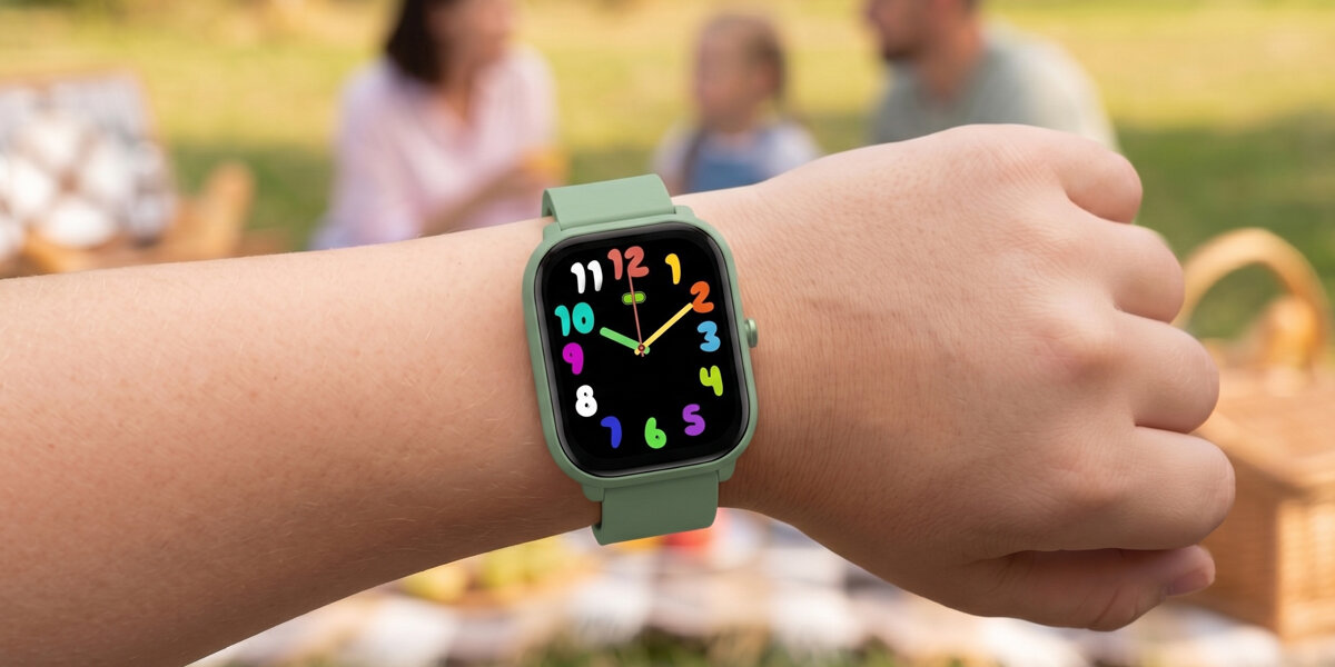 Na zdjęciu widoczny jest zielony smartwatch z kolorowymi cyframi na tarczy, umieszczony na nadgarstku osoby. W tle rozmyty obraz pikniku z ludźmi.