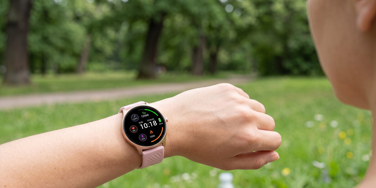 Kobieta patrzy na różowy smartwatch Denver z okrągłą tarczą. W tle znajduje się rozmyty, zielony park.