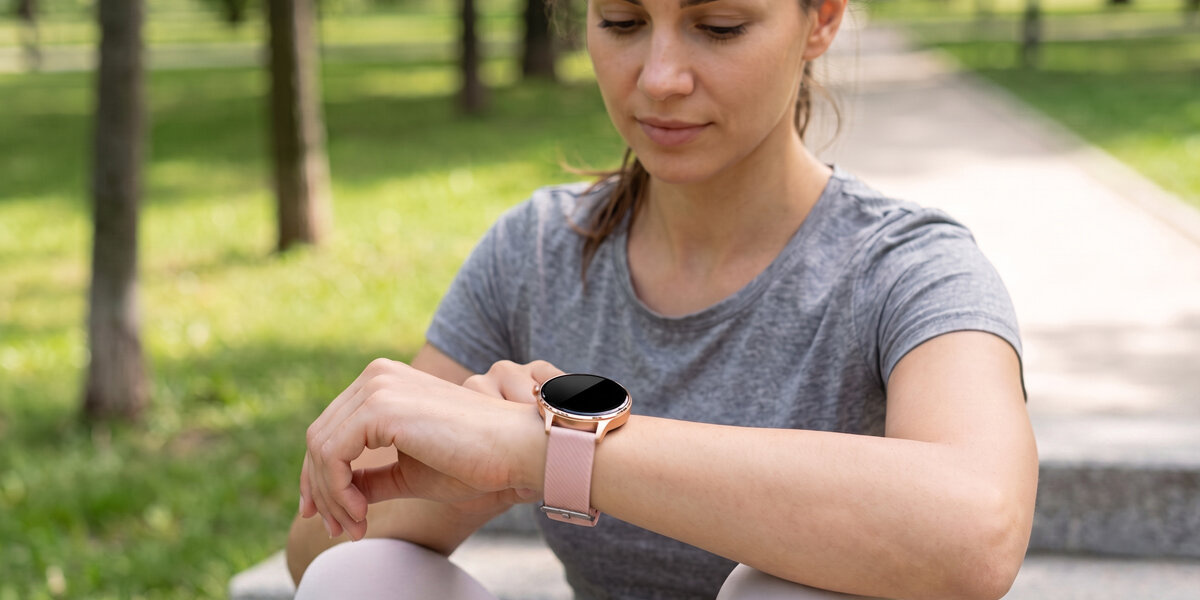 Kobieta w szarej koszulce siedzi na schodach. Na jej ręce znajduje się różowy smartwatch z wygaszonym ekranem.