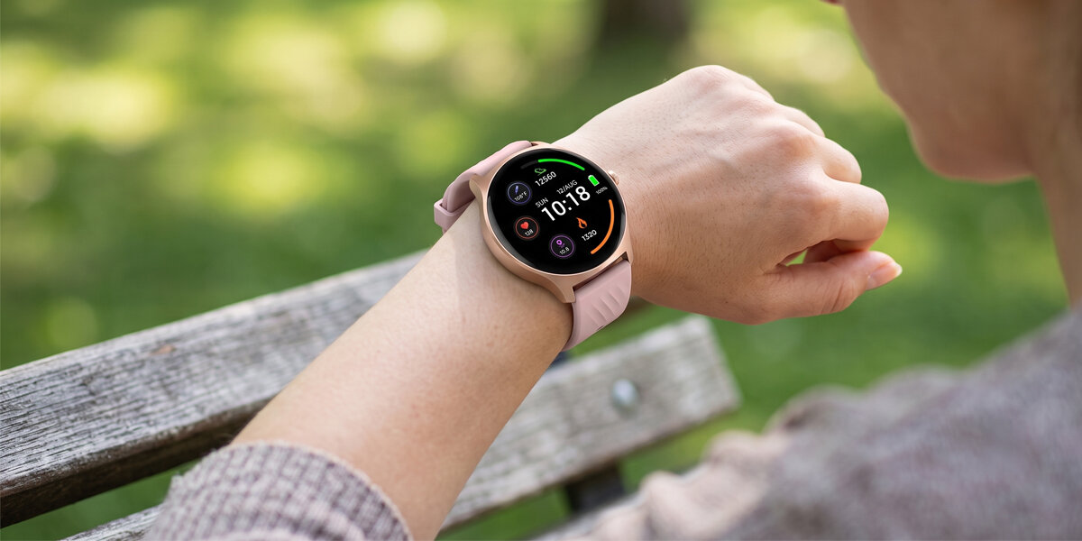 Kobieta patrzy na różowy smartwatch Denver, stojąc przed ławką. Wokół znajduje się pełen zieleni park.