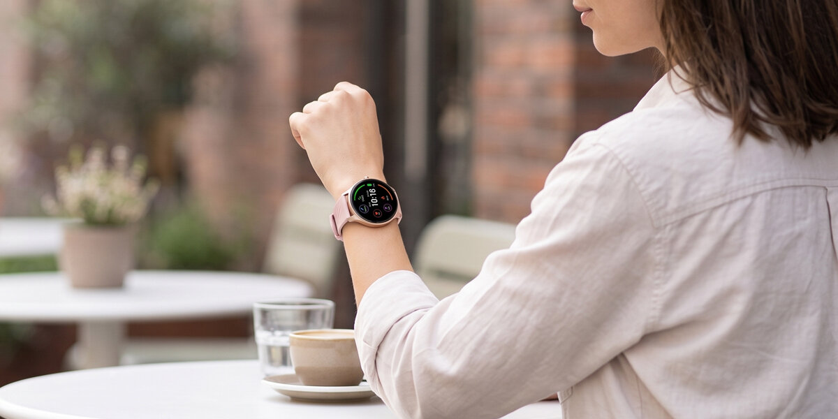 Kobieta siedzi w kawiarnianym ogródku. Na jej ręce znajduje się różowy, okrągły smartwatch Denver z włączonym ekranem.