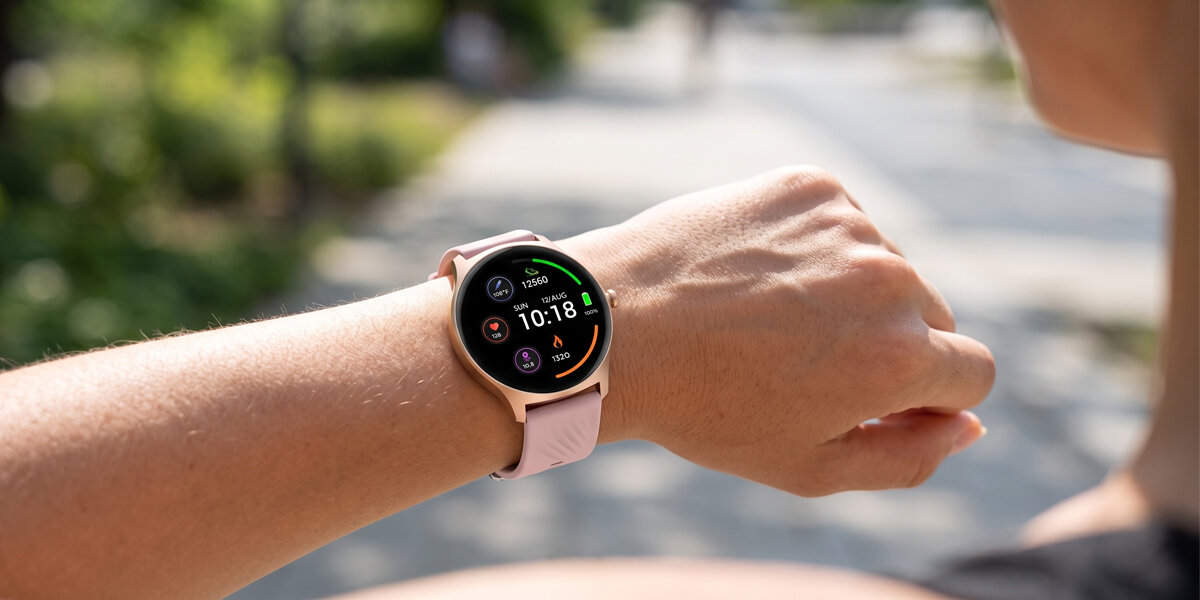 Na wysuniętym przed siebie nadgarstku kobiety znajduje się różowy smartwatch Denver. W zamazanym tle jest ścieżka w parku.