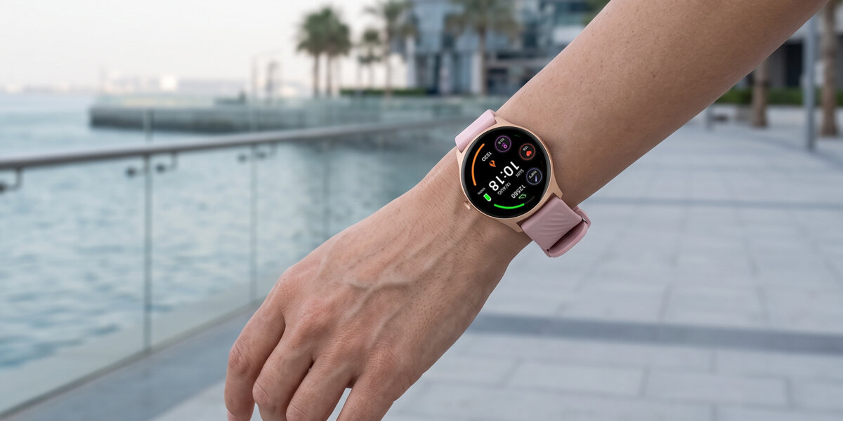 Na wysuniętej ręce znajduje się różowy smartwatch Denver. W tle widać nadmorską promenadę z palmami.