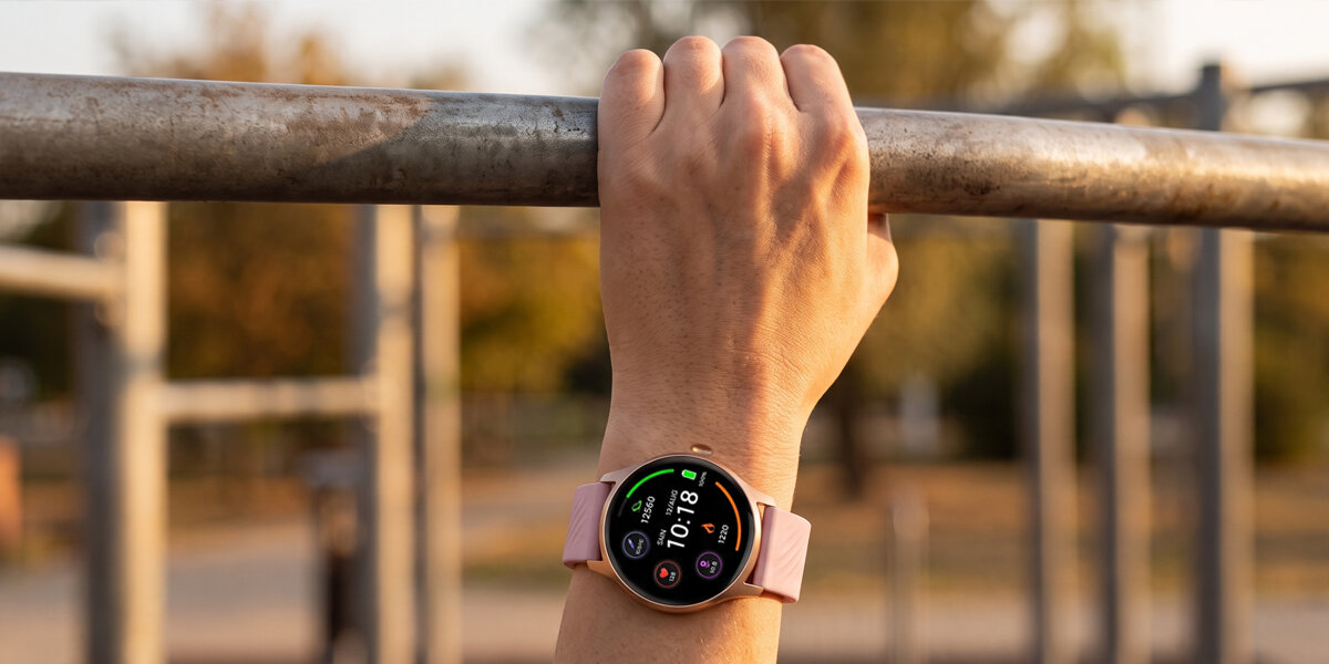Dłoń zaciska się na metalowym drążku do ćwiczeń. Na nadgarstku znajduje się różowy smartwatch z jasnym ekranem.