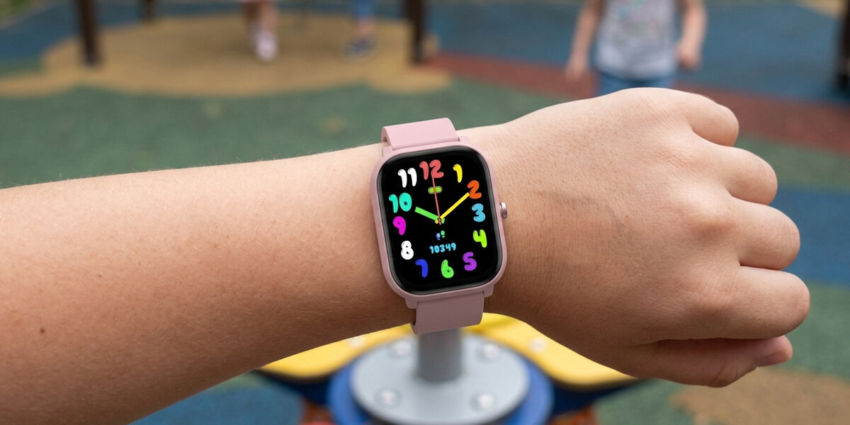 Osoba nosi różowy smartwatch z kolorowym wyświetlaczem, na którym widoczne są cyfry od 1 do 12 w różnych kolorach oraz napis \'10:34\'.