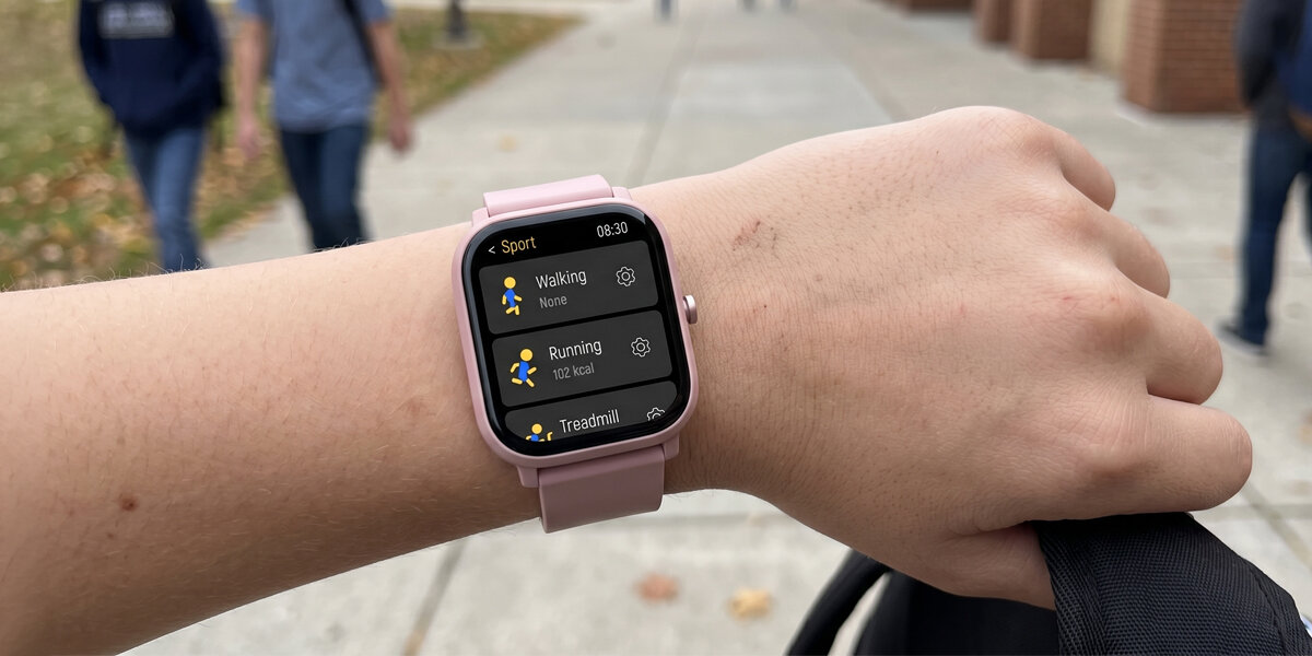 Na zdjęciu widoczny jest różowy smartwatch na nadgarstku, z wyświetlaczem pokazującym opcje sportowe: Walking, Running (102 kcal) i Treadmill. W tle widać rozmyte postacie i chodnik.