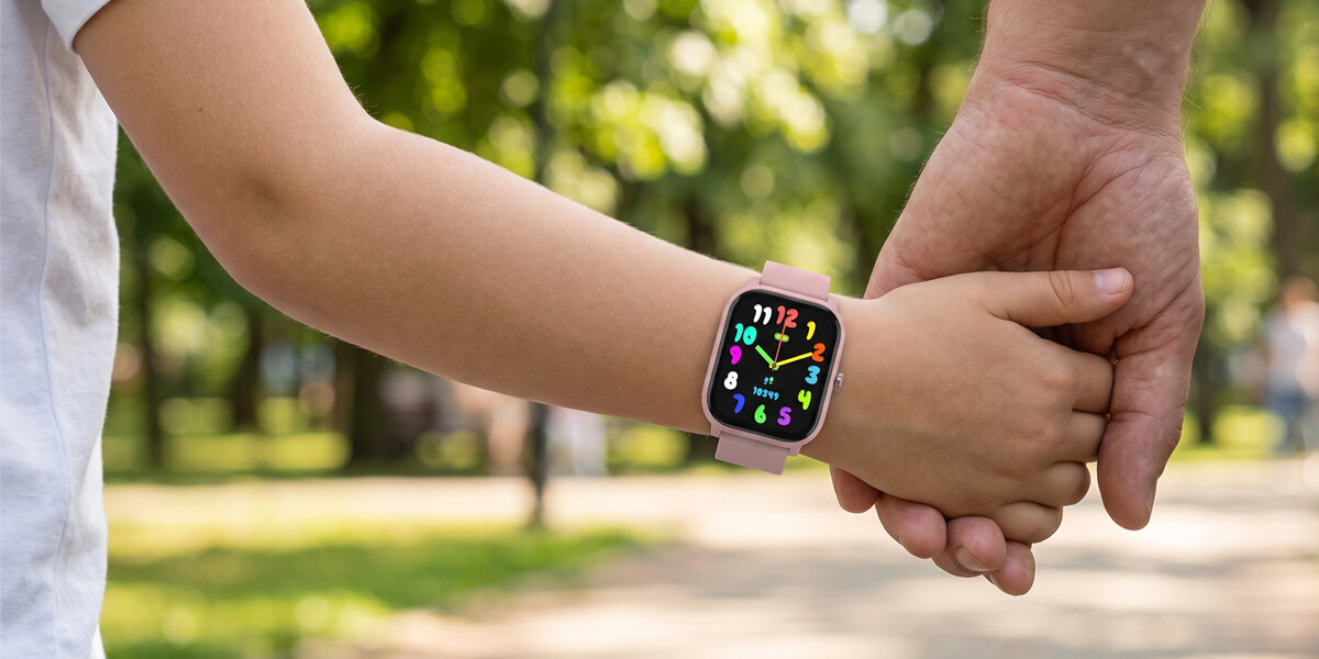 Dziecko nosi różowy smartwatch z kolorowym wyświetlaczem, trzymając za rękę dorosłą osobę. W tle widać rozmyty park z zielenią.