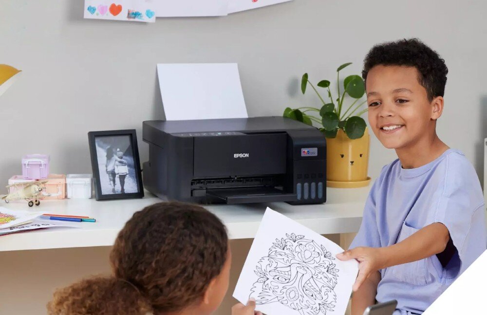Na biurku stoi urządzenie wielofunkcyjne EPSON z widocznym logo, obok którego znajduje się roślina w doniczce i ramka ze zdjęciem. Dwoje dzieci siedzi przy biurku, jedno z nich trzyma kartkę z kolorowanką.