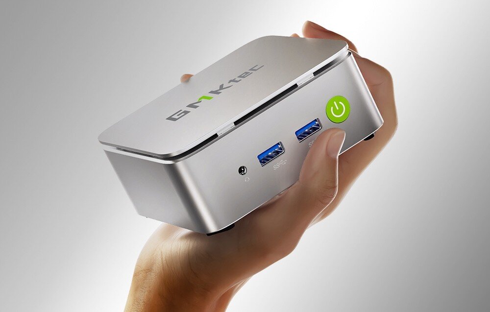Komputer GMKTEC G10 Osoba trzyma w dłoni mały srebrny komputer z widocznymi dwoma portami USB i zielonym przyciskiem z symbolem włączenia, obudowa, wielkość