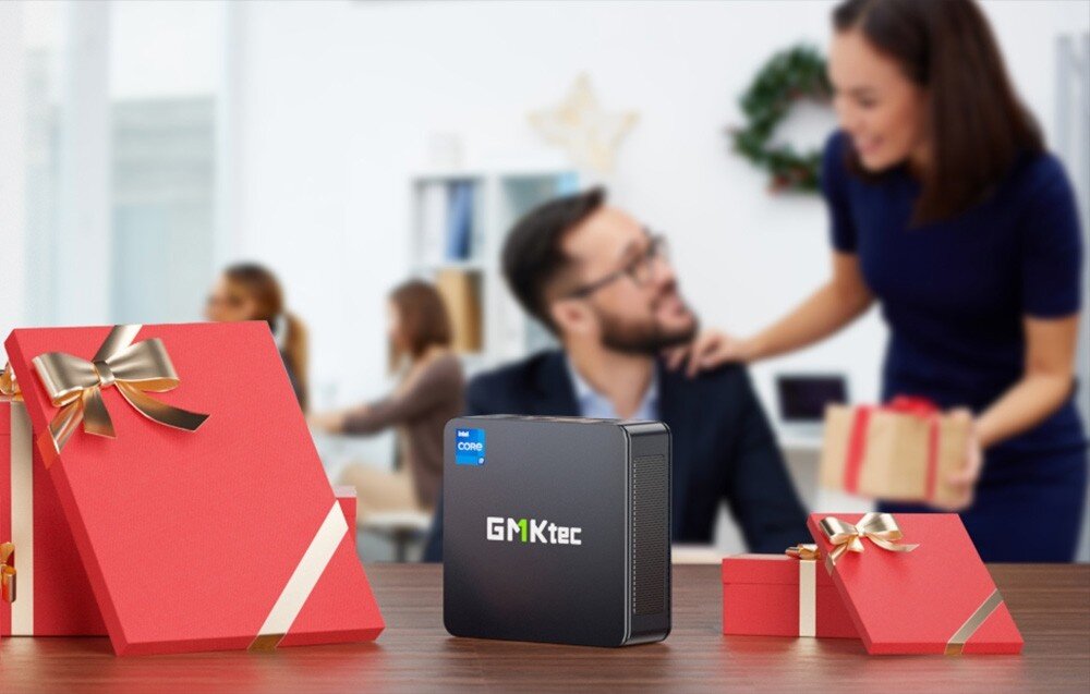 Komputer GMKTEC K7 Plus Mały czarny komputer ustawiony na biurku obok czerwonych pudełek prezentowych, w tle kobieta wręcza mężczyźnie upominek, opis