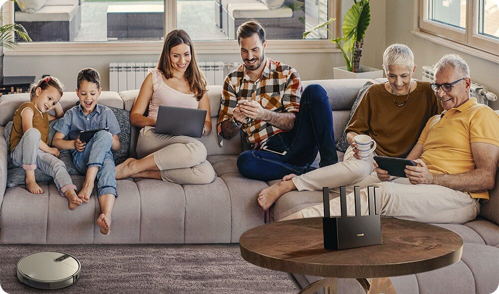 Na kanapie siedzi grupa osób korzystających z różnych urządzeń elektronicznych. Na stoliku przed nimi znajduje się router z widocznym logo \'tenda\'.