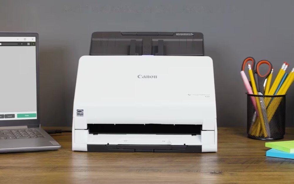 Skaner CANON R30 Skaner w kolorze białym ustawiony na drewnianym blacie obok otwartego laptopa, przyborów piśmienniczych w czarnym przyborniku oraz kolorowych karteczek samoprzylepnych, opis