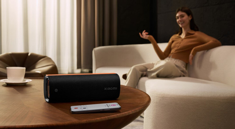 Głośnik mobilny XIAOMI Sound Party, IPX5, akumulator, mobilność, osoba siedząca na sofie, głośnik leżący na stoliku obok smartfona
