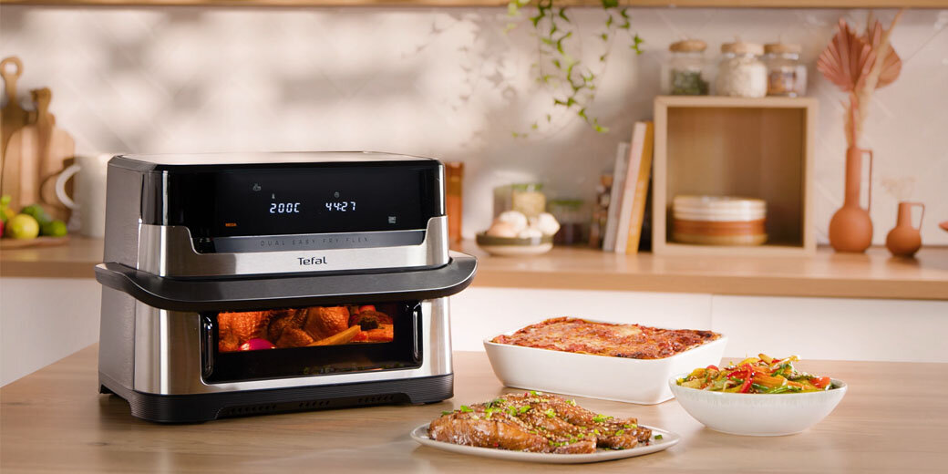 Frytkownica Dual Easy Fry Flex  EY922D na blacie kuchennym, w jej wnętrzu piecze się kurczak. Obok gotowe potrawy.