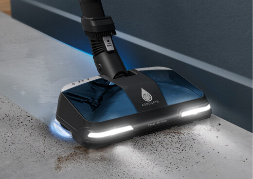 Odkurzacz z funkcją mopowania czyszczący podłogę, z widocznymi światłami LED i logo \'AEROSPIN\'. Na podłodze widać zabrudzenia, które są usuwane przez urządzenie.