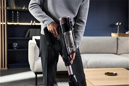 Osoba trzyma odkurzacz pionowy z przezroczystym pojemnikiem na kurz w nowoczesnym wnętrzu z sofą i stolikiem. Odkurzacz ma metaliczny różowy element i czarną rączkę.