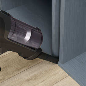 Odkurzacz z elastyczną rurą czyszczący podłogę przy krawędzi mebla. Podłoga jest drewniana, a mebel ma szary kolor.