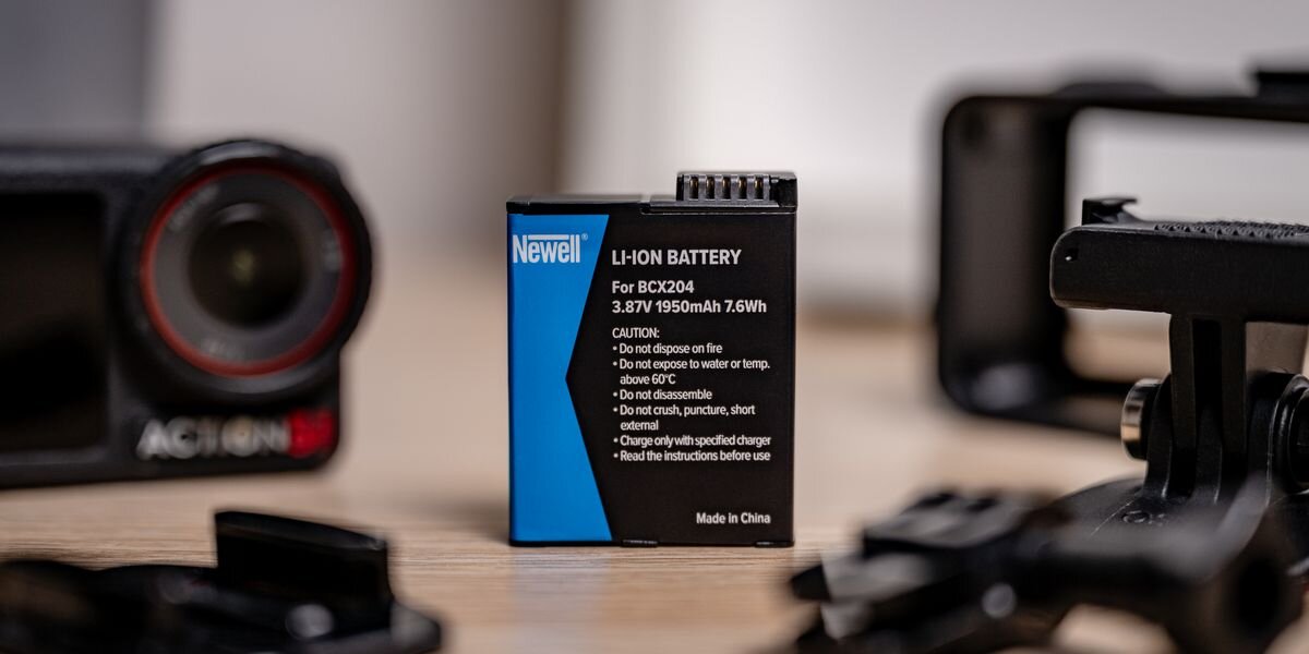 Akumulator NEWELL zamiennik BCX204 do DJI Osmo Action 3/4/5 Pro, Akumulator Newell ustawiony pionowo na jasnym blacie, w tle rozmyta kamera sportowa oraz czarna ramka obudowy, dokładne, ładowarka, elastyczny, obudowa