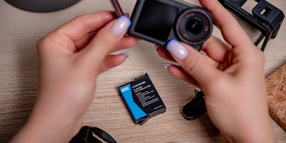 Akumulator NEWELL zamiennik BCX204 do DJI Osmo Action 3/4/5 Pro, Dłonie trzymające kamerę sportową z otwartą klapką, obok na blacie leży czarno-niebieski akumulator marki Newell, IC, przegrzanie, przeciążenie, przeładowanie