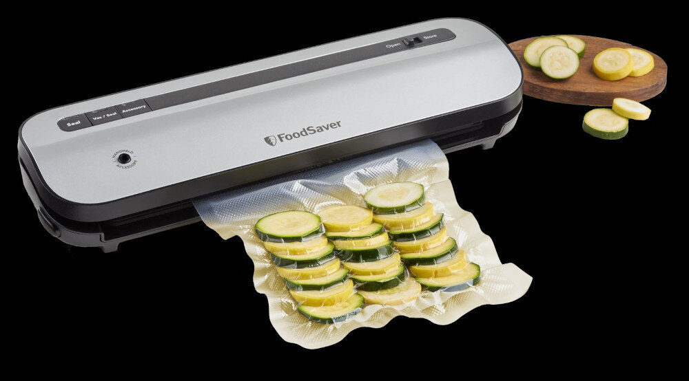 Zgrzewarka próżniowa z logo \'FoodSaver\' na obudowie, z zapakowanymi w folię plasterkami cukinii. Obok urządzenia znajduje się deska z dodatkowymi plasterkami cukinii i żółtej cukinii.