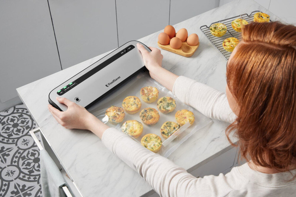 Osoba używa zgrzewarki próżniowej marki FoodSaver do pakowania jedzenia w przezroczystą folię. Na blacie obok znajdują się jajka i wypieki na metalowej kratce.