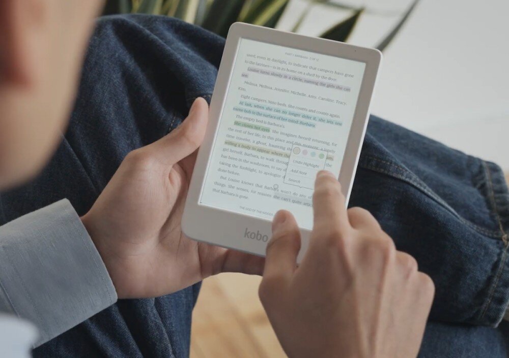 Czytnik e-booków KOBO Clara Colour Osoba siedząca z nogą założoną na nogę używa białego czytnika e-booków, dotykając palcem ekranu z wyświetlonym tekstem, notatki, organizacja, podkreślanie