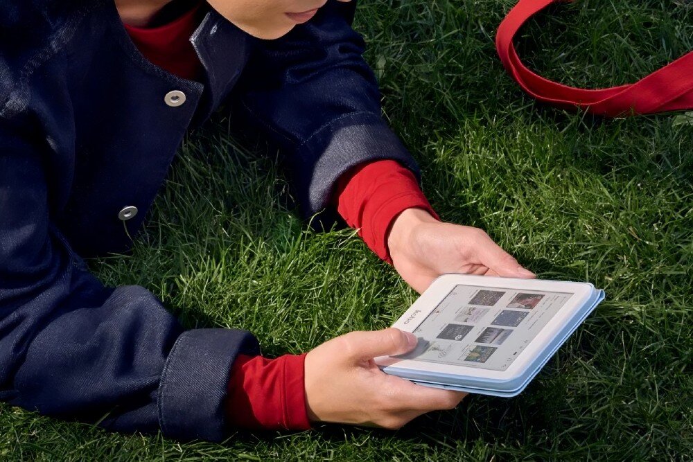 Czytnik e-booków KOBO Clara Colour Osoba leży na trawie w plenerze i trzyma biały czytnik e-booków w dłoniach, na ekranie widoczne są miniatury książek, pamięć, bateria, czas działania, ładowanie