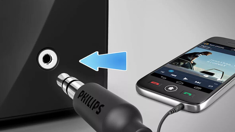 Obraz przedstawia zbliżenie na gniazdo audio oraz wtyczkę z napisem \'PHILIPS\', obok której znajduje się strzałka wskazująca na gniazdo. Obok widoczny jest smartfon z odtwarzaczem muzyki na ekranie.