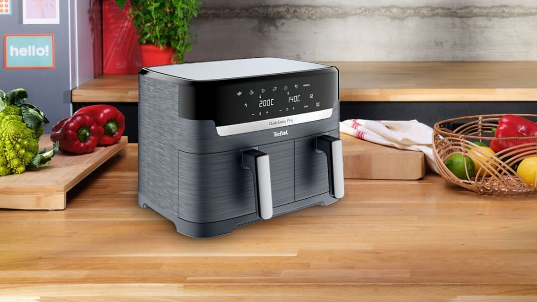 Na blacie kuchennym stoi frytkownica beztłuszczowa z wyświetlaczem pokazującym temperatury 200°C i 140°C. Obok znajdują się warzywa i koszyk z owocami, a na urządzeniu widnieje logo \'Tefal\'.