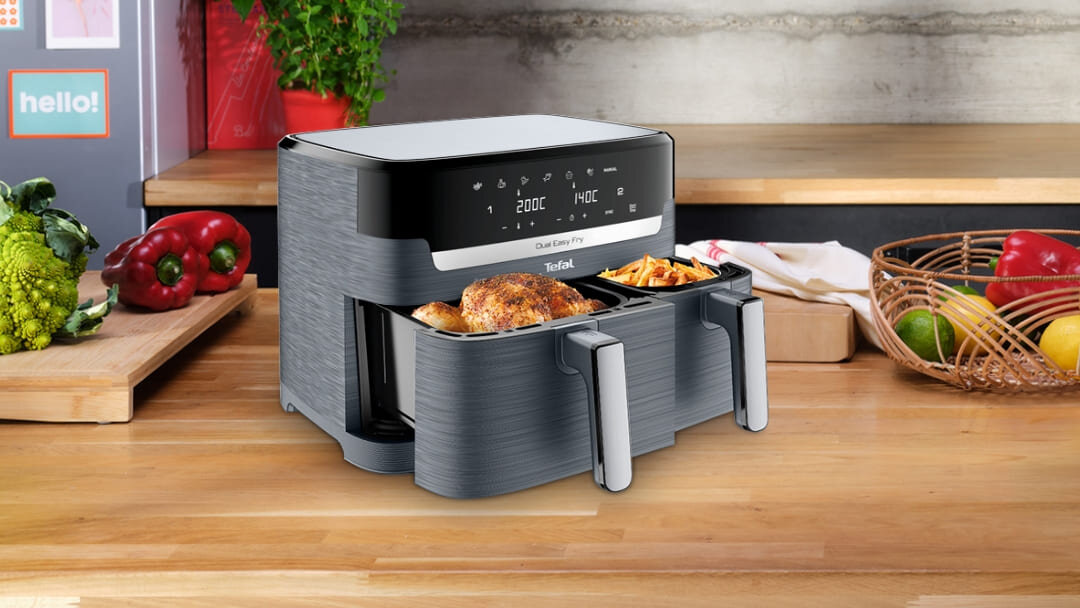 Na blacie kuchennym stoi frytkownica beztłuszczowa z dwoma wysuniętymi koszami, w których znajdują się pieczony kurczak i frytki. Na panelu dotykowym urządzenia widoczne są ustawienia temperatury 200°C i czasu 10:06.