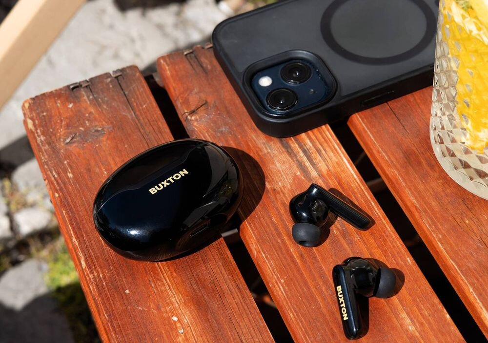Słuchawki dokanałowe BUXTON BTW 7200 Tuya IPX6 wodoodporność 5,5 h pracy szybkie ładowanie USB-C etui 25 h, słuchawki z etui ładującym oraz smartfonem leżące na stoliku