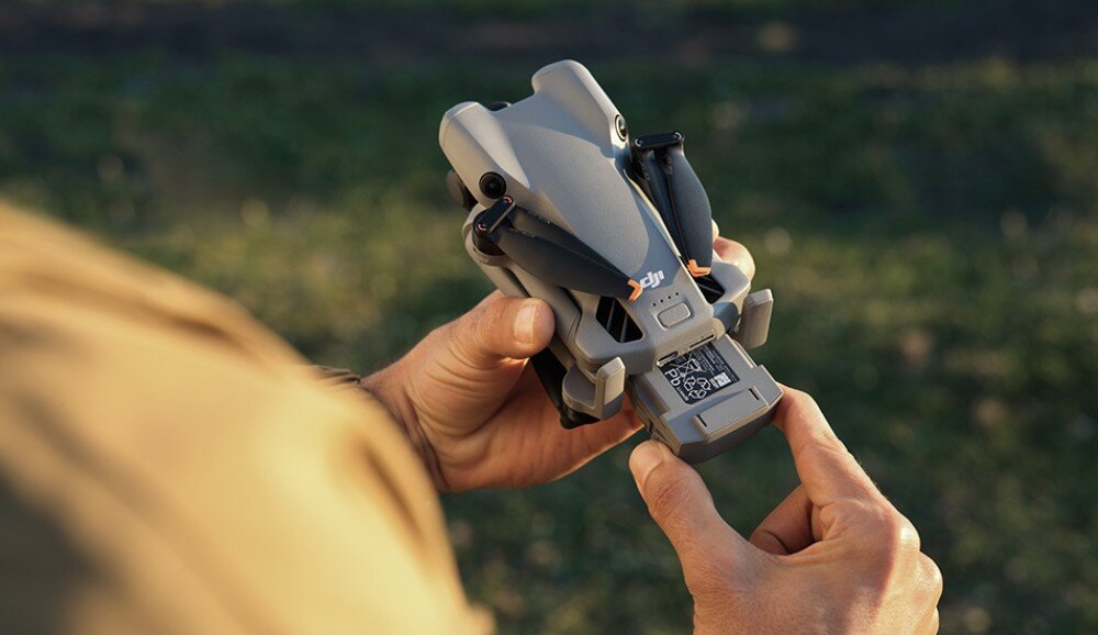 Dron DJI Mini 5 Pro Zbliżenie na ręce trzymające złożonego drona, w którym wysuwany jest akumulator z kodem QR, czas lotu, ładowanie, zasięg, prędkość