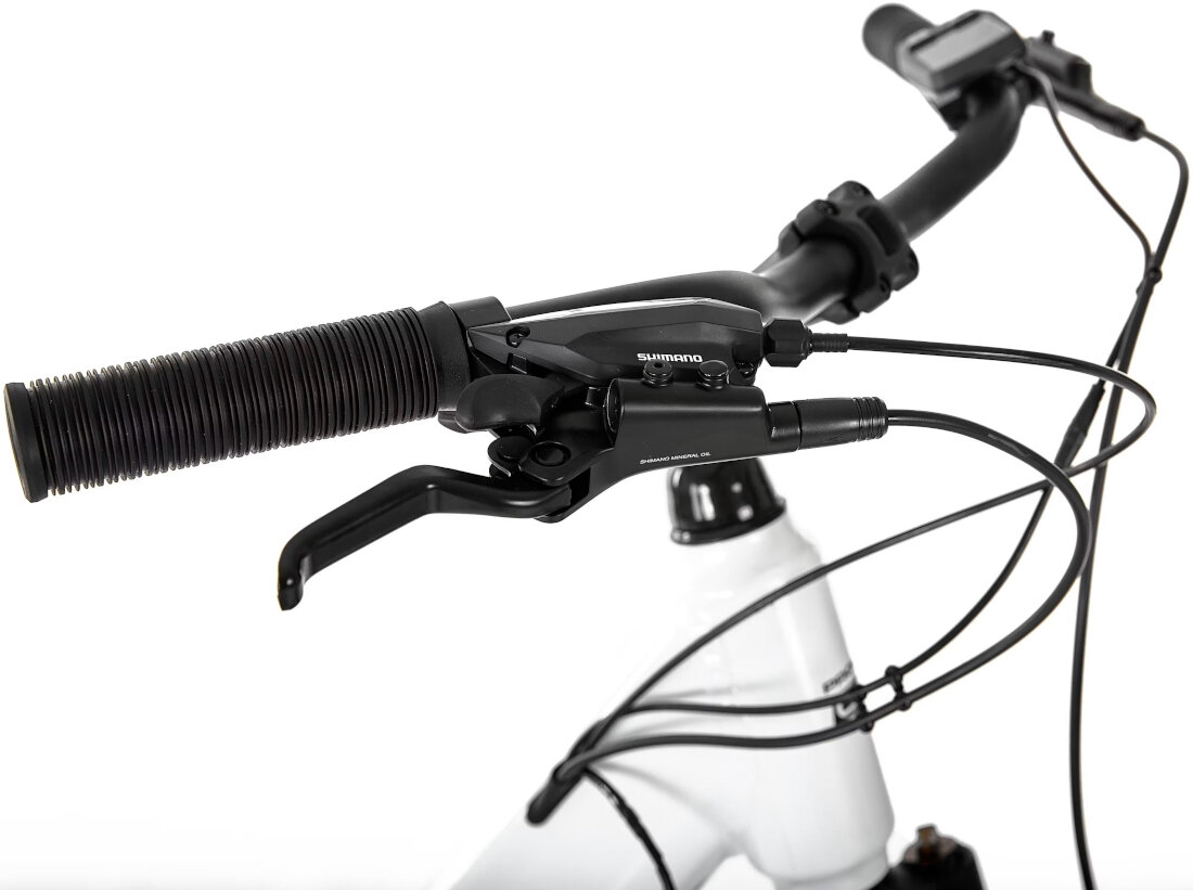 Zbliżenie na kierownicę roweru z widocznym czarnym uchwytem i dźwignią hamulca z napisem \'Shimano\'. W tle widać fragment białej ramy roweru oraz przewody hamulcowe.