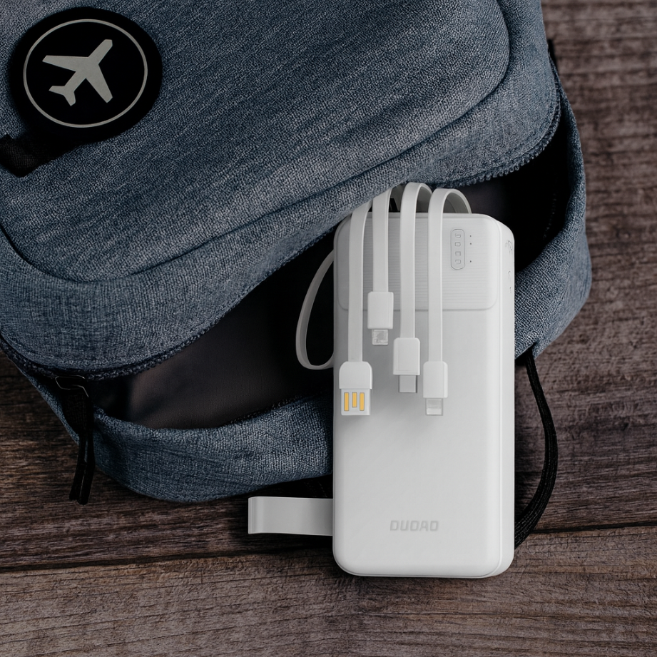 Biały powerbank z wbudowanymi kablami leży obok niebieskiego plecaka. Na plecaku widoczna jest naszywka z symbolem samolotu.