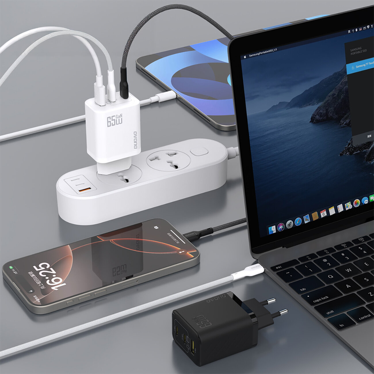 Na obrazie widoczna jest ładowarka sieciowa z napisem \'65W GAN\' podłączona do listwy zasilającej, obok której znajdują się smartfon i laptop. Obok leży czarna ładowarka z napisem \'65W GAN\'.