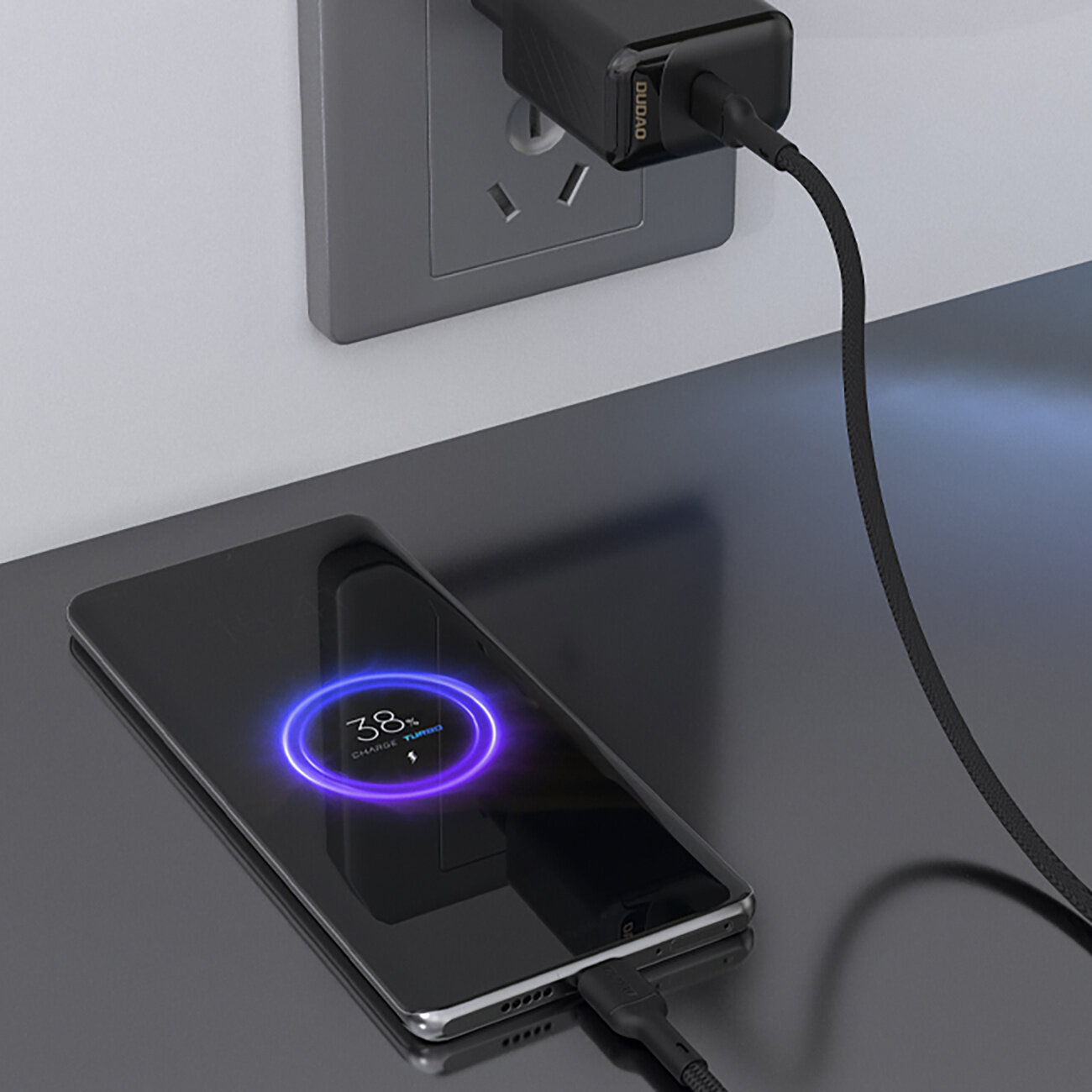 Telefon leży na powierzchni, podłączony do ładowarki za pomocą czarnego kabla USB-C. Na ekranie telefonu widoczny jest wskaźnik ładowania z napisem \'38%\'.