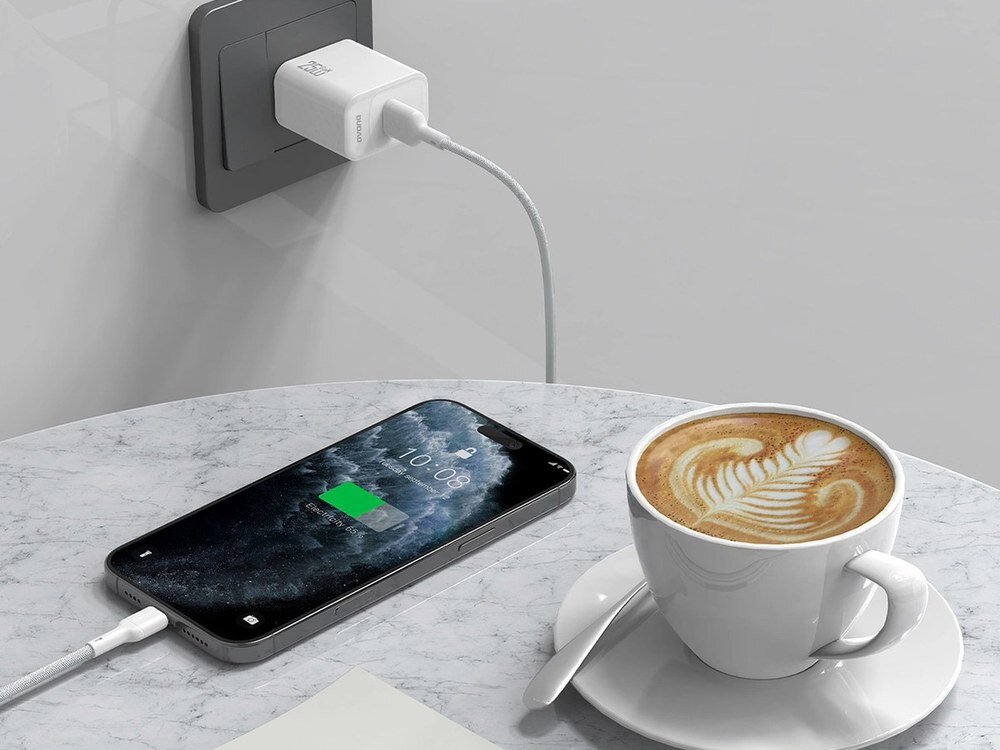 Na marmurowym stole leży smartfon podłączony do ładowarki w gniazdku ściennym za pomocą białego kabla USB-C. Obok znajduje się filiżanka kawy z wzorem na piance.