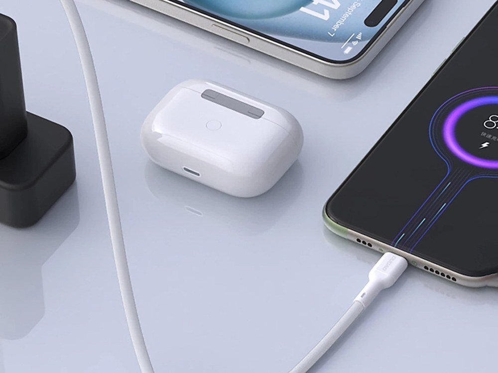 Na obrazie widoczny jest biały kabel USB-C podłączony do smartfona, obok którego znajduje się etui na słuchawki bezprzewodowe. W tle widać fragment innego smartfona oraz czarną ładowarkę.