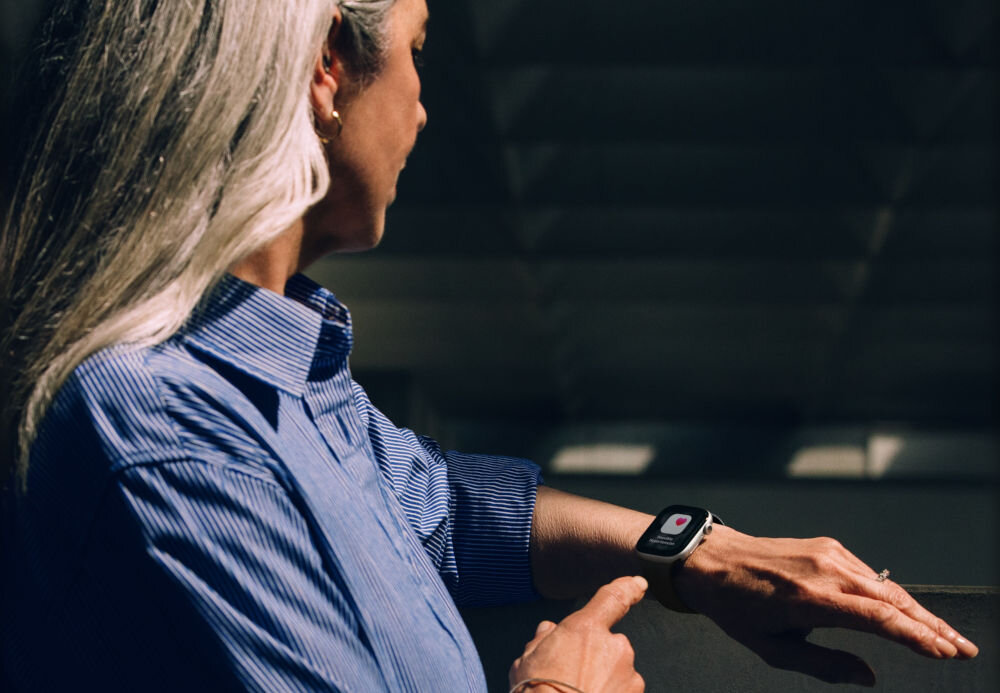 Apple Watch 11 GPS 42mm koperta z aluminium (różowe złoto) + pasek sportowy rozmiar S/M (łagodny róż) kobieta w niebieskiej koszuli z zegarkiem na ręku pokazująca palcem na niego w tle rozmazany dach budynku funkcja powiadomień o nadciśnieniu monitorowanie zdrowia badania kliniczne analiza danych z optycznego czujnika tętna